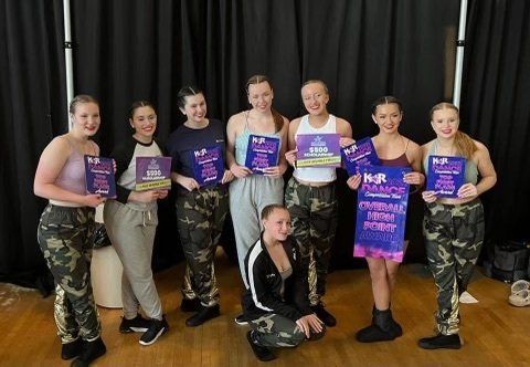 A group of performers holding purple awards and plaques in front of black curtains, posing on a stage