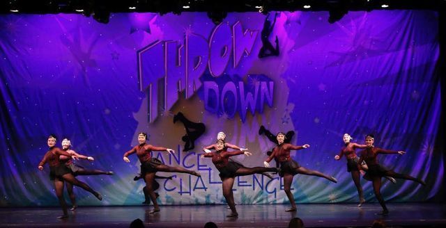 Dancers in black costumes perform on a purple-lit stage with a “Throw Down” backdrop.