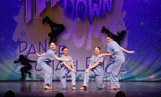 Five dancers in gray costumes perform on a purple-lit stage with 