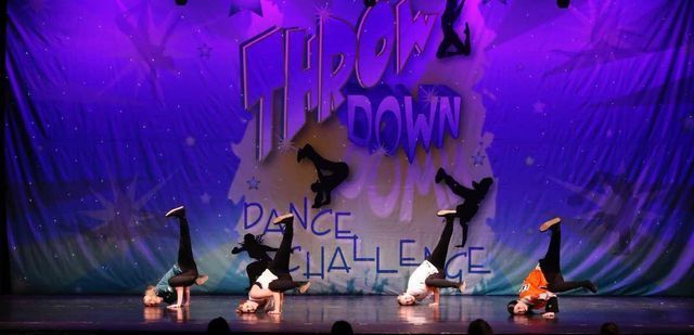 Four dancers in black perform on a purple-lit stage with a large graffiti-style backdrop.