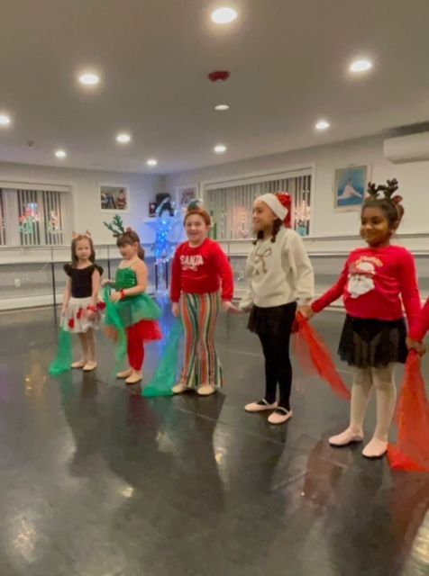 Children in a dance studio wearing holiday costumes, standing in a line with colorful tulle skirts and Santa hat.