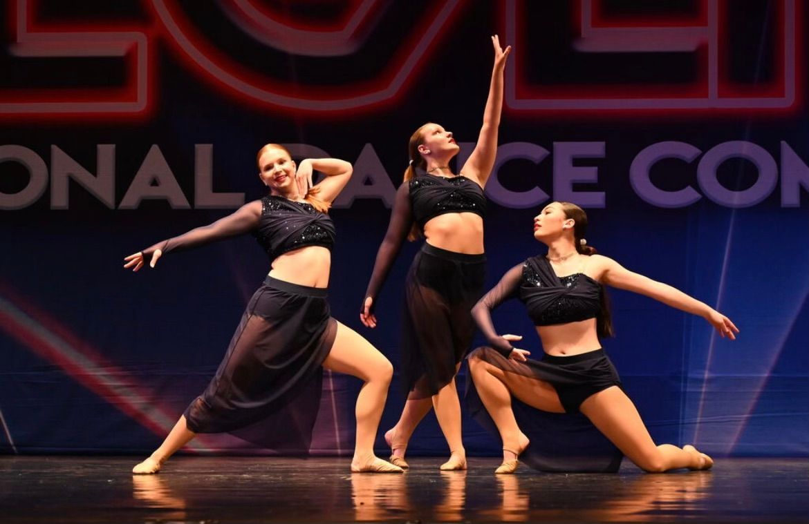 Three dancers in black costumes perform on a stage with a purple-lit backdrop.
