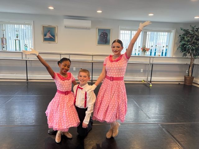 Three dancers in pink costumes posing in a ballet studio, two with arms raised.