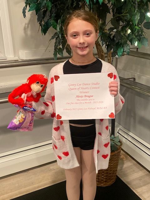 Girl holding a handwritten sign beside a red-haired doll, wearing a heart-patterned cardigan indoors.