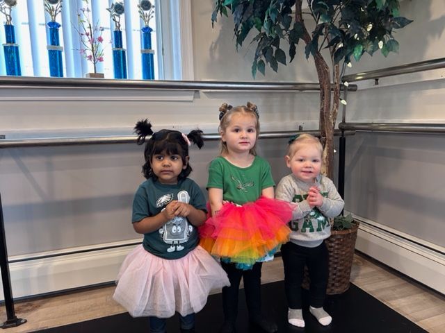 Three children in colorful tutus standing indoors, posing by a railing and wall.