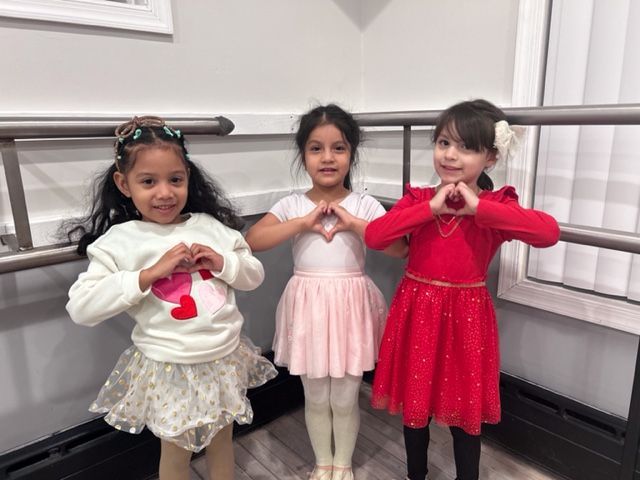 Three children in ballet outfits posing with hands in heart shapes at a barre in a dance studio