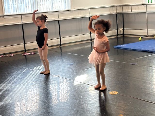 Two children in ballet outfits standing in a dance studio with arms raised