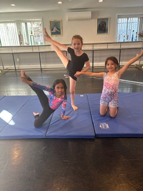 Three girls posing in dance costumes on blue mats in a studio, arms and legs extended.