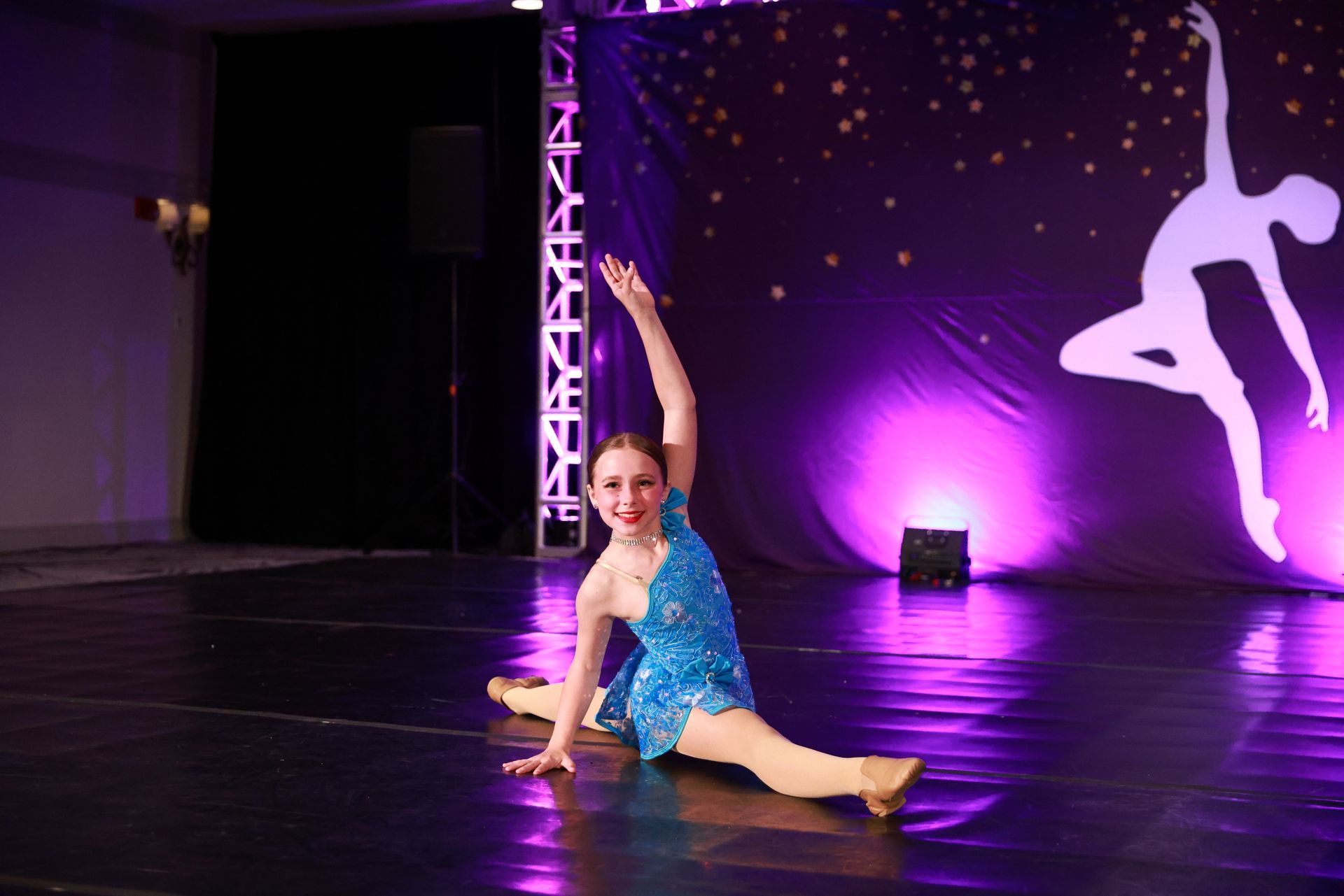 Young dancer in blue costume doing a split on a stage with purple lighting and a lit backdrop