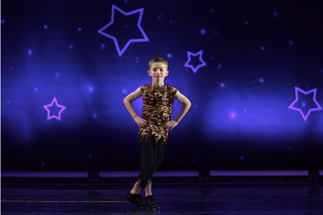 Young dancer in a gold costume posing on a purple-lit stage with star decorations.