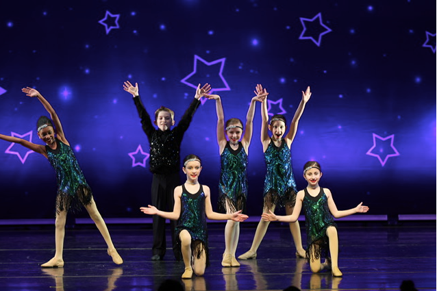 Six dancers in green costumes perform on a purple stage with starry lights behind them