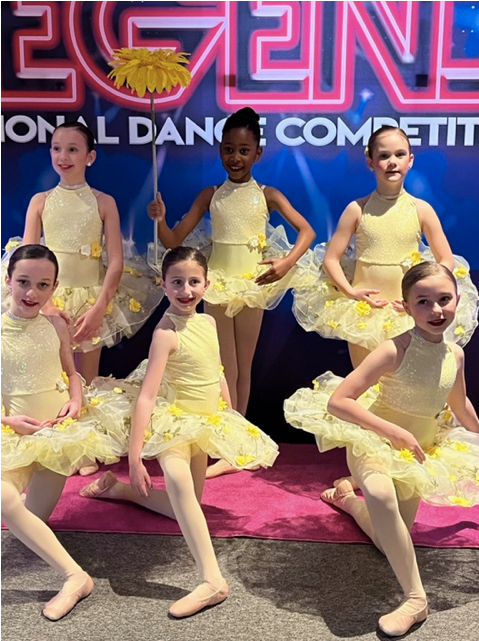 Six young dancers in yellow costumes posing on stage in front of a dance competition backdrop