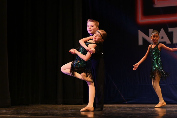 Two dancers in black sequined dresses perform a lift on a stage, with another dancer in the background.