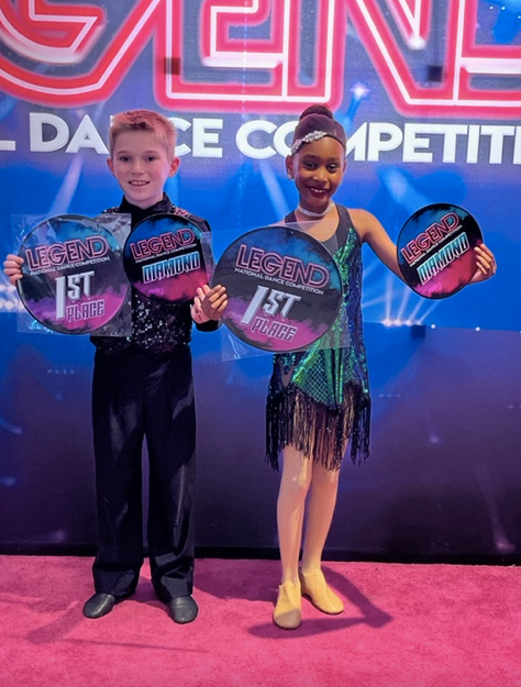 Two dancers holding 1st place awards at a dance competition stage.
