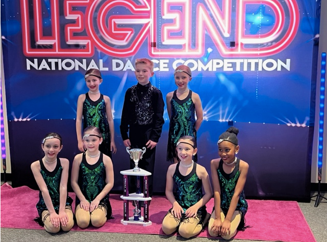 Dance team posing with trophy on pink carpet under “LEGEND National Dance Competition” sign