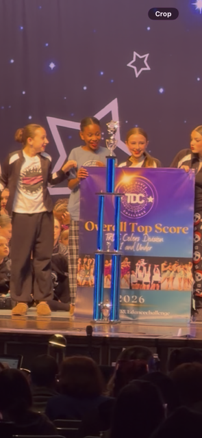 Three children on stage beside a purple podium, holding a trophy under starry lights.