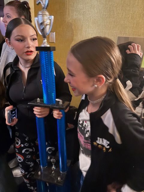 Three girls at a party holding a large blue trophy, one in a black jacket smiling at the camera