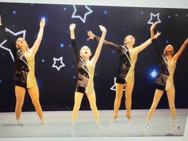 Five dancers in gold and black leotards pose with arms raised on a starry stage.