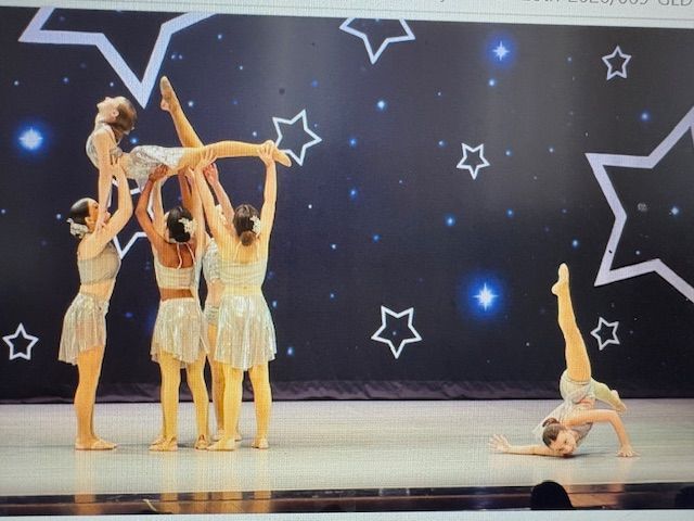 Four dancers in silver costumes pose on stage against a starry backdrop, with two balancing in lifts.