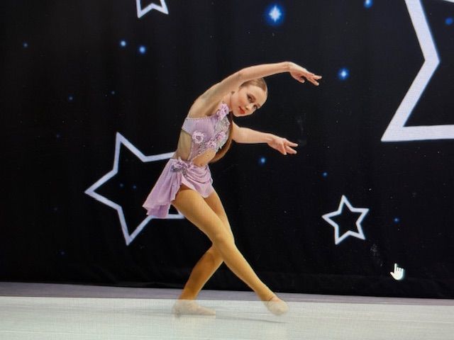 A young dancer in a pink costume poses onstage against a black starry backdrop.