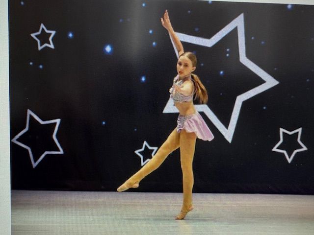 Young dancer posing onstage in a pink-and-white costume against a black star backdrop.