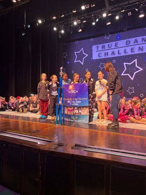Children on a stage holding a large trophy at a dance challenge event, with purple lights and star backdrop.