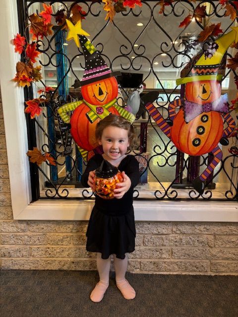 Smiling child in black dress holding an orange treat, standing before colorful Halloween decorations.