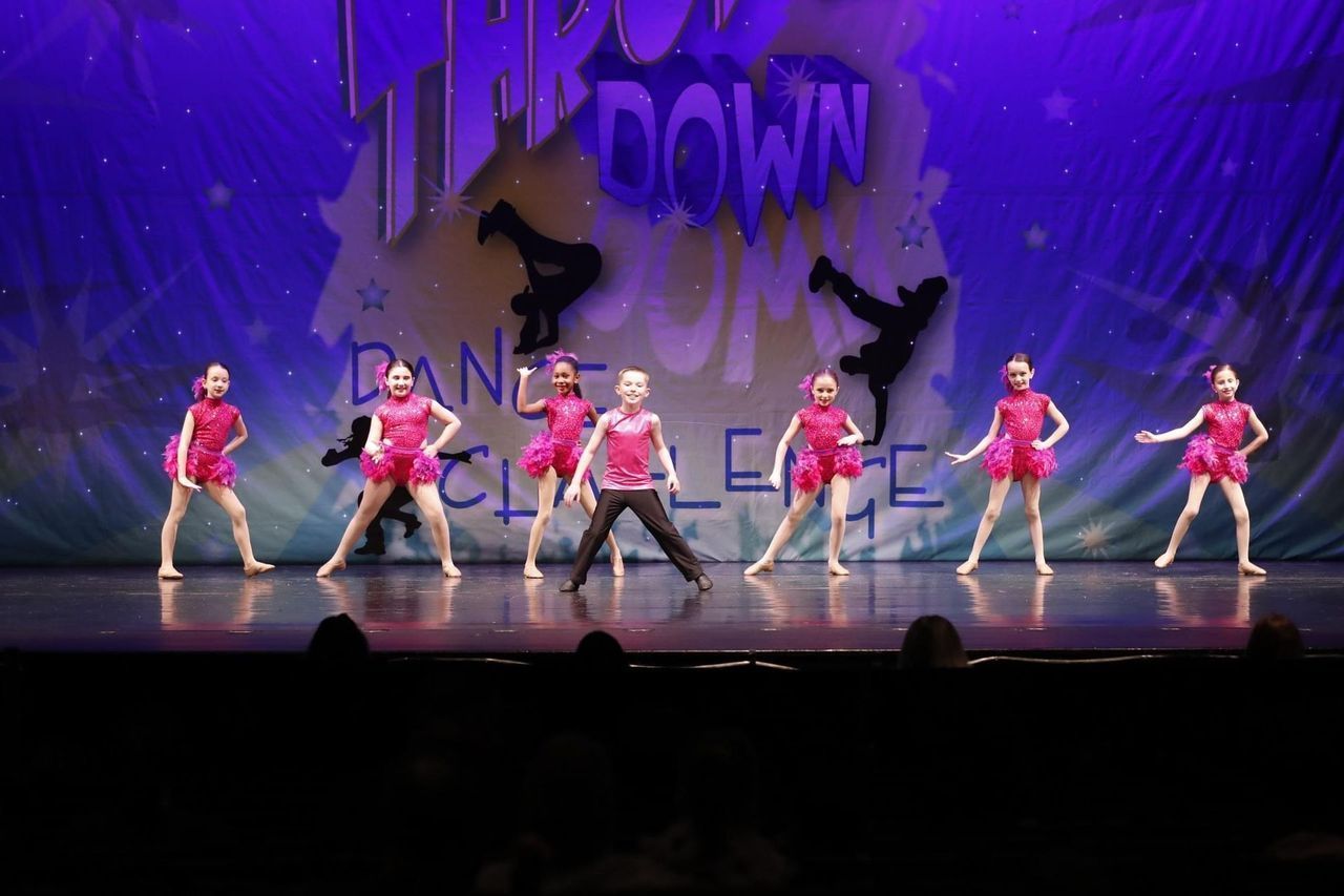 Seven dancers in pink perform on a lit stage with a purple backdrop and shadowy wolves.