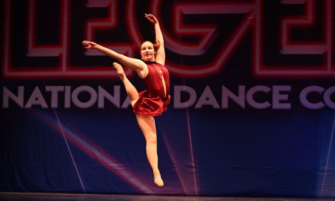 Dancer in red costume performing a high leg extension on stage at National Dance Competition
