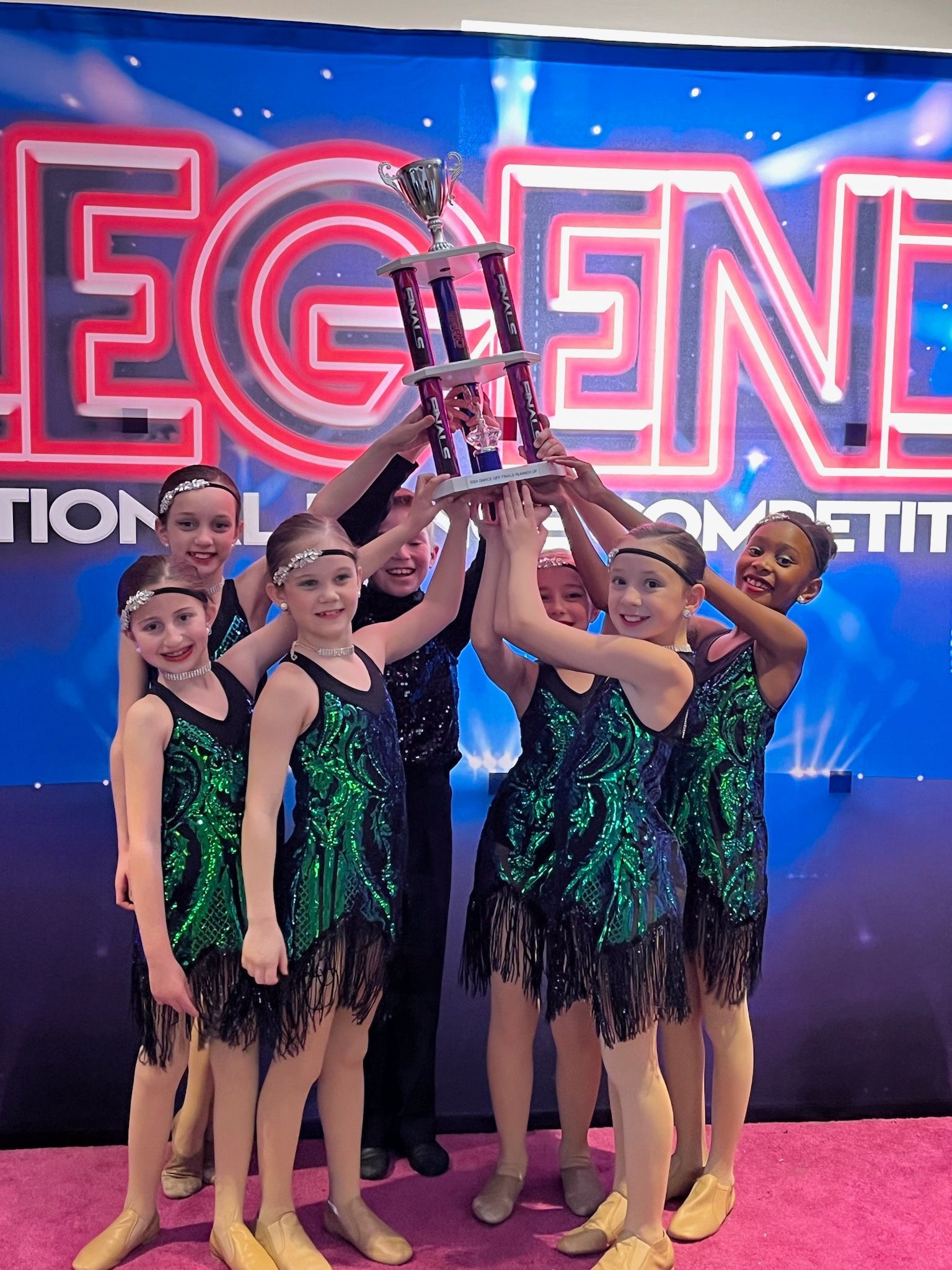 Dance team holding a trophy onstage in green-and-black costumes, celebrating a competition win.