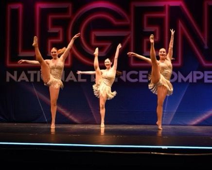 Three dancers in gold costumes striking high-leg poses on a lit competition stage