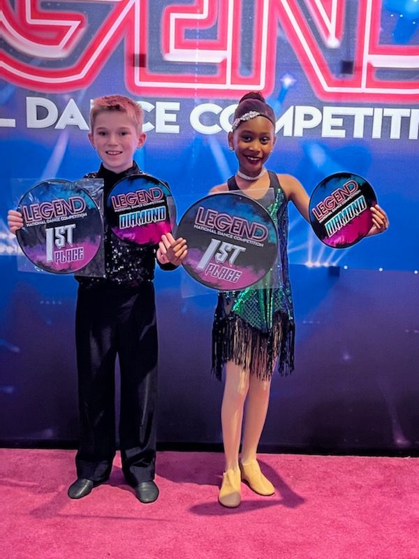 Two dance competition winners holding first-place awards on a stage with a neon backdrop