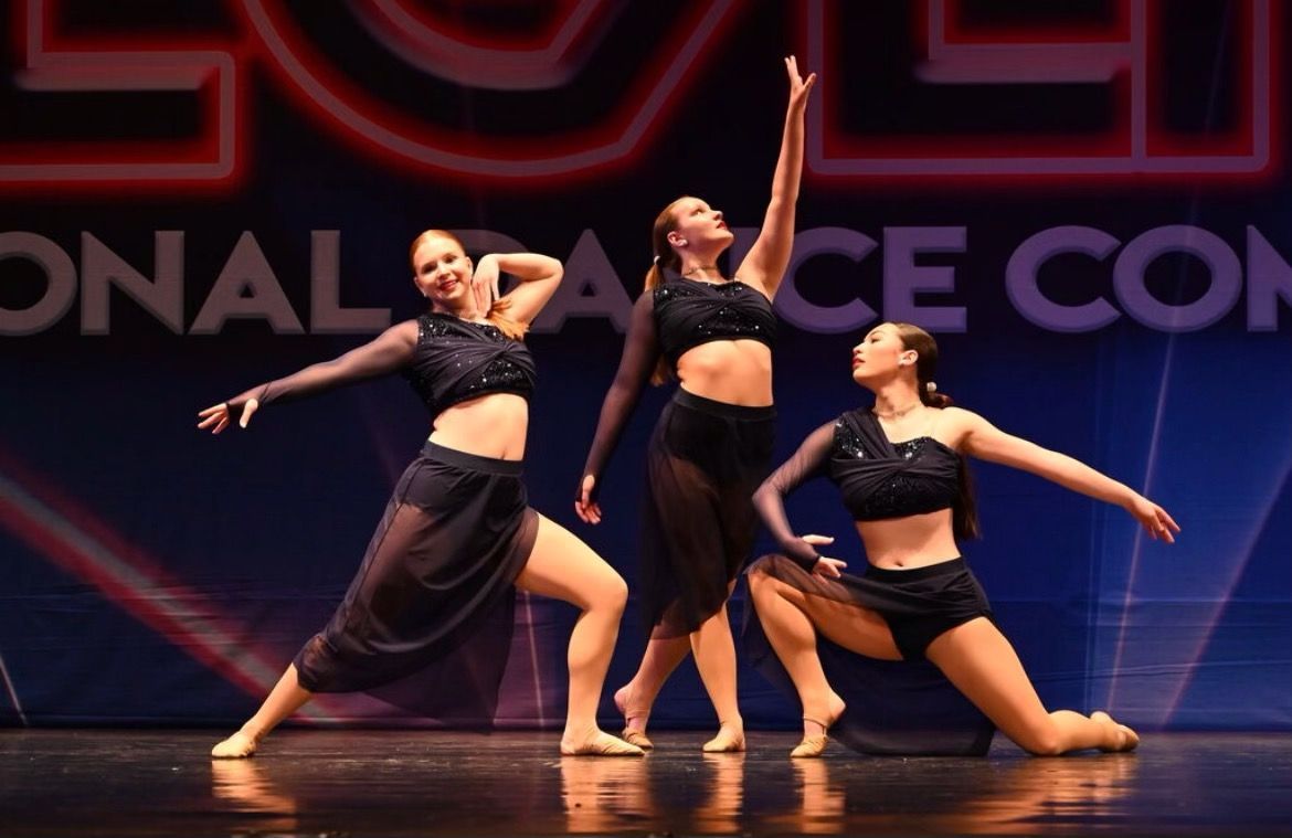 Three dancers in black costumes strike a dramatic pose on a stage during a competition