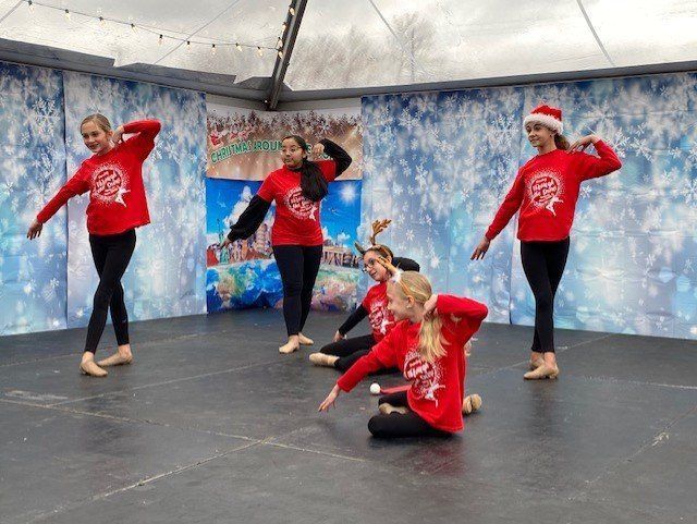 Four dancers in red holiday sweaters pose on an outdoor stage with a snowy backdrop.