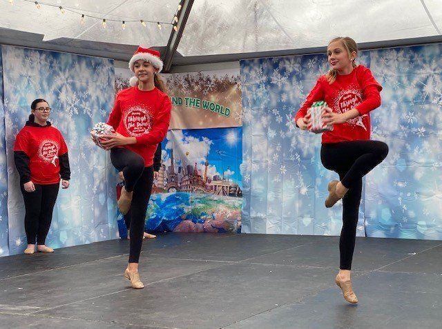 Three dancers in red holiday tops balancing on one leg on an indoor stage with a snowy backdrop