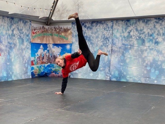 Breakdancer in a red shirt doing a one-handed handstand on a stage with a blue backdrop