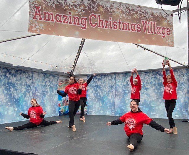 Five dancers in red shirts pose on an outdoor stage under an “Amazing Christmas Village” banner.