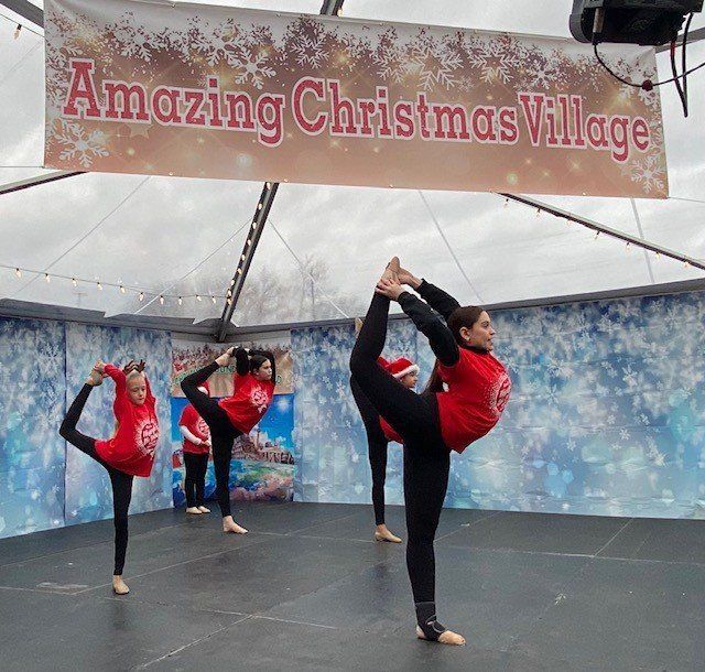 Three dancers in red and black pose on stage under an “Amazing Christmas Village” banner.