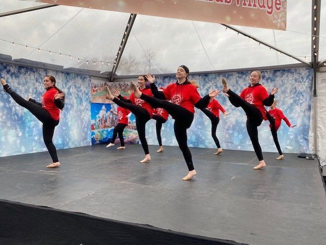 Five dancers in red tops and black pants perform high kicks on a stage under a tent