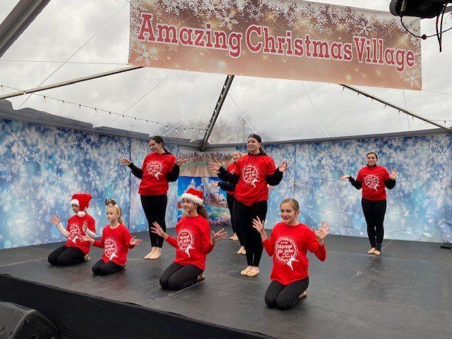 Children in red shirts performing a dance on an outdoor stage under an “Amazing Christmas Village” banner