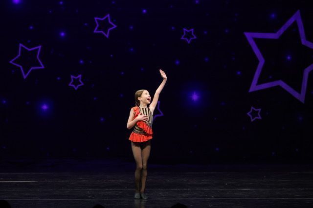 Dancer in a red costume on a dark stage, reaching upward amid purple star graphics