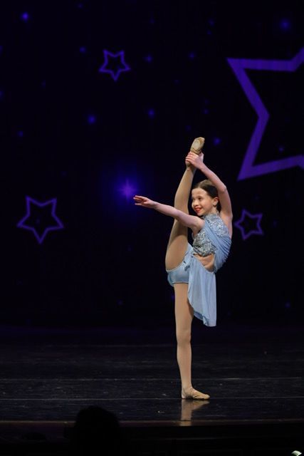 Young dancer in a blue costume performs a high leg hold on a dark stage with purple star decorations