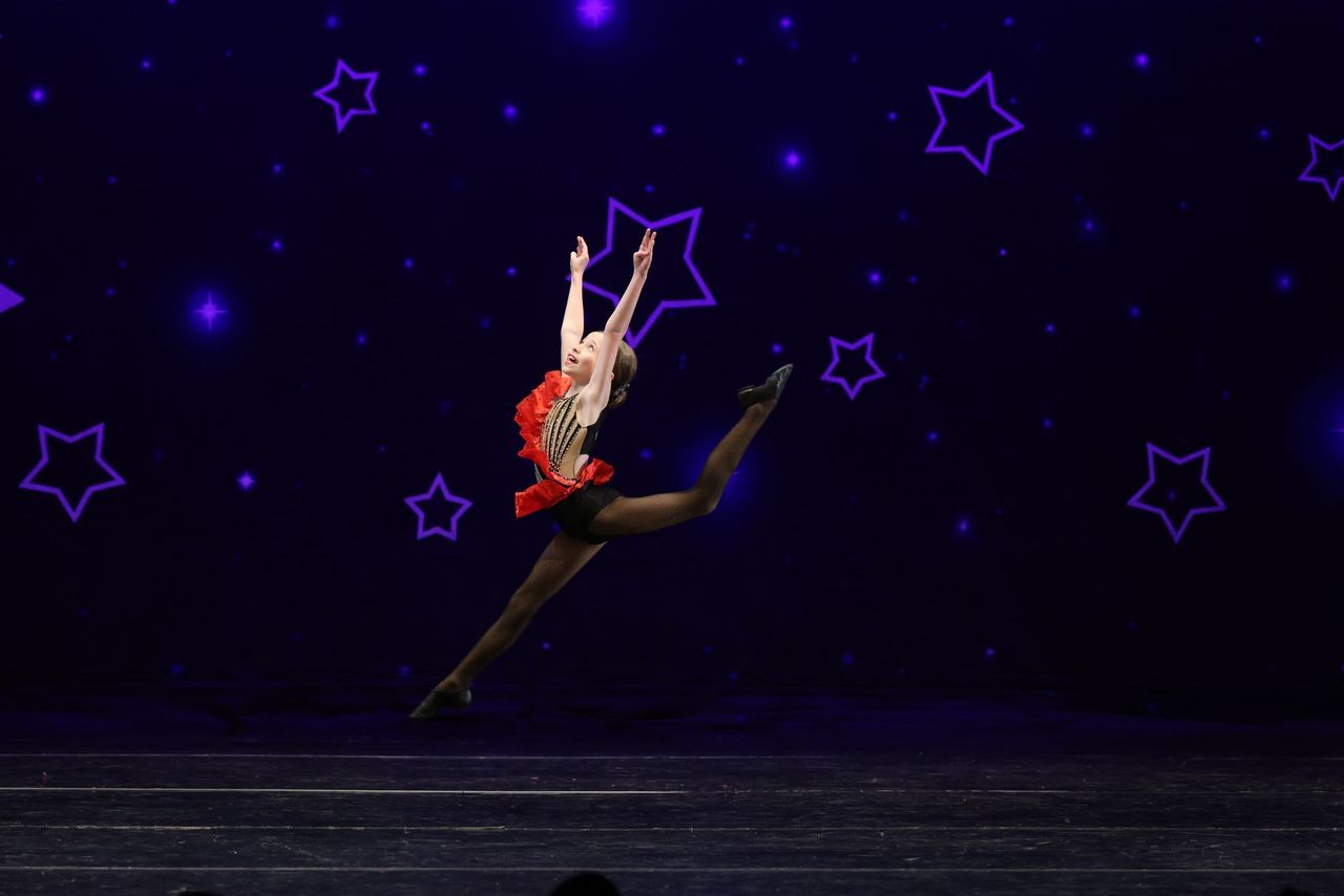 Dancer in red and black leaps midair on a dark stage with glowing purple stars in the background