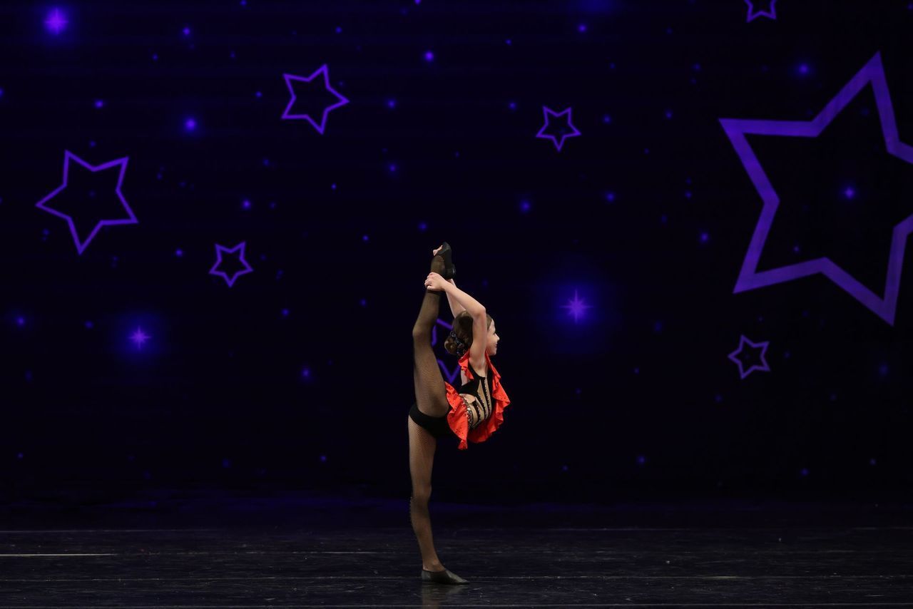 Contortion dancer in red costume balancing on one leg against a dark stage with purple star lights