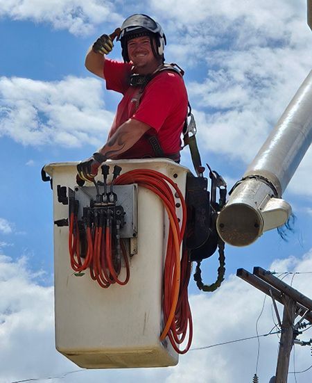 Utility worker in a bucket lift, wearing helmet and red shirt, high above the ground against a cloudy sky.