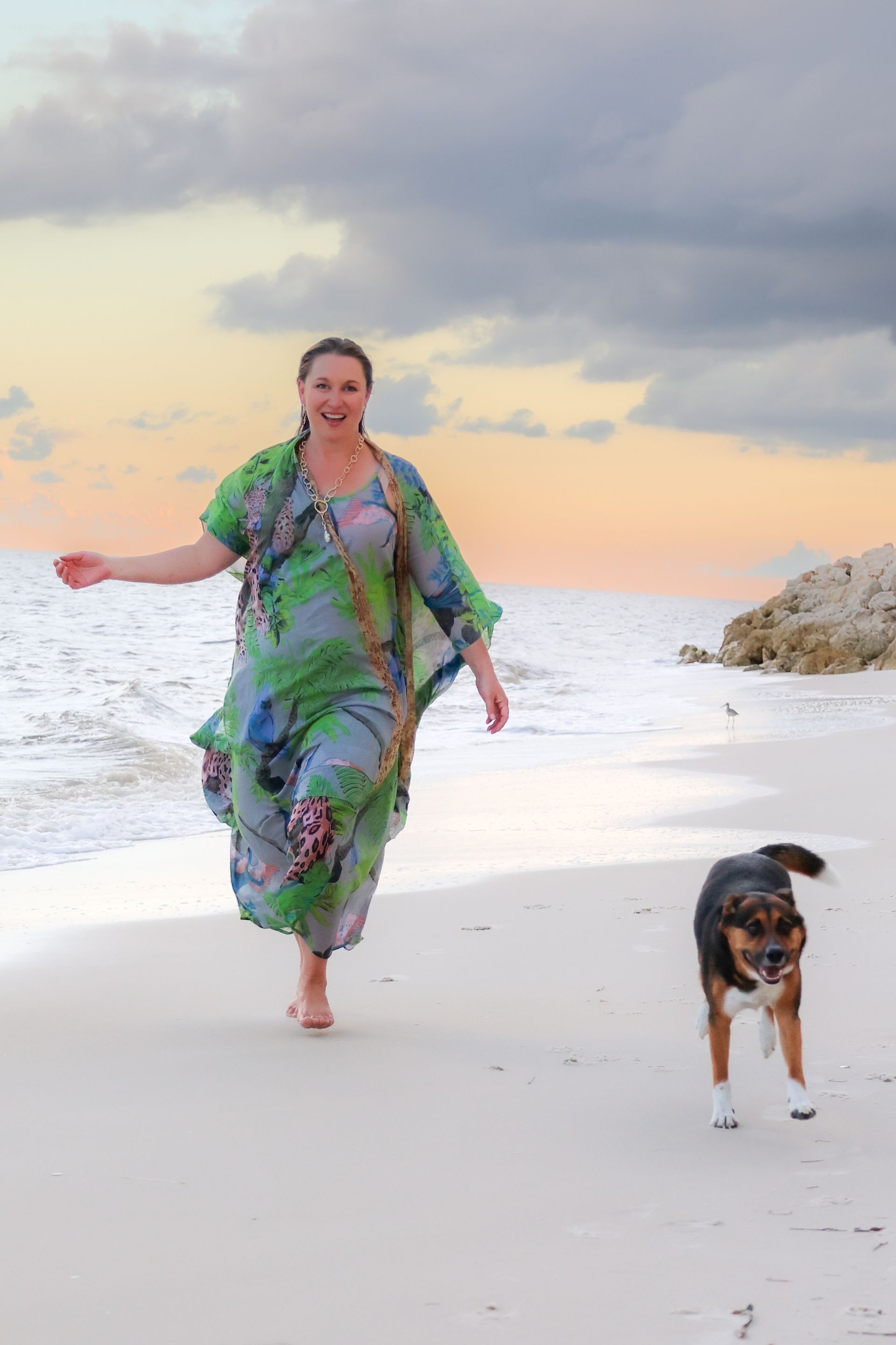 Woman in a flowing dress runs on beach with a dog; sunset sky and ocean in background.