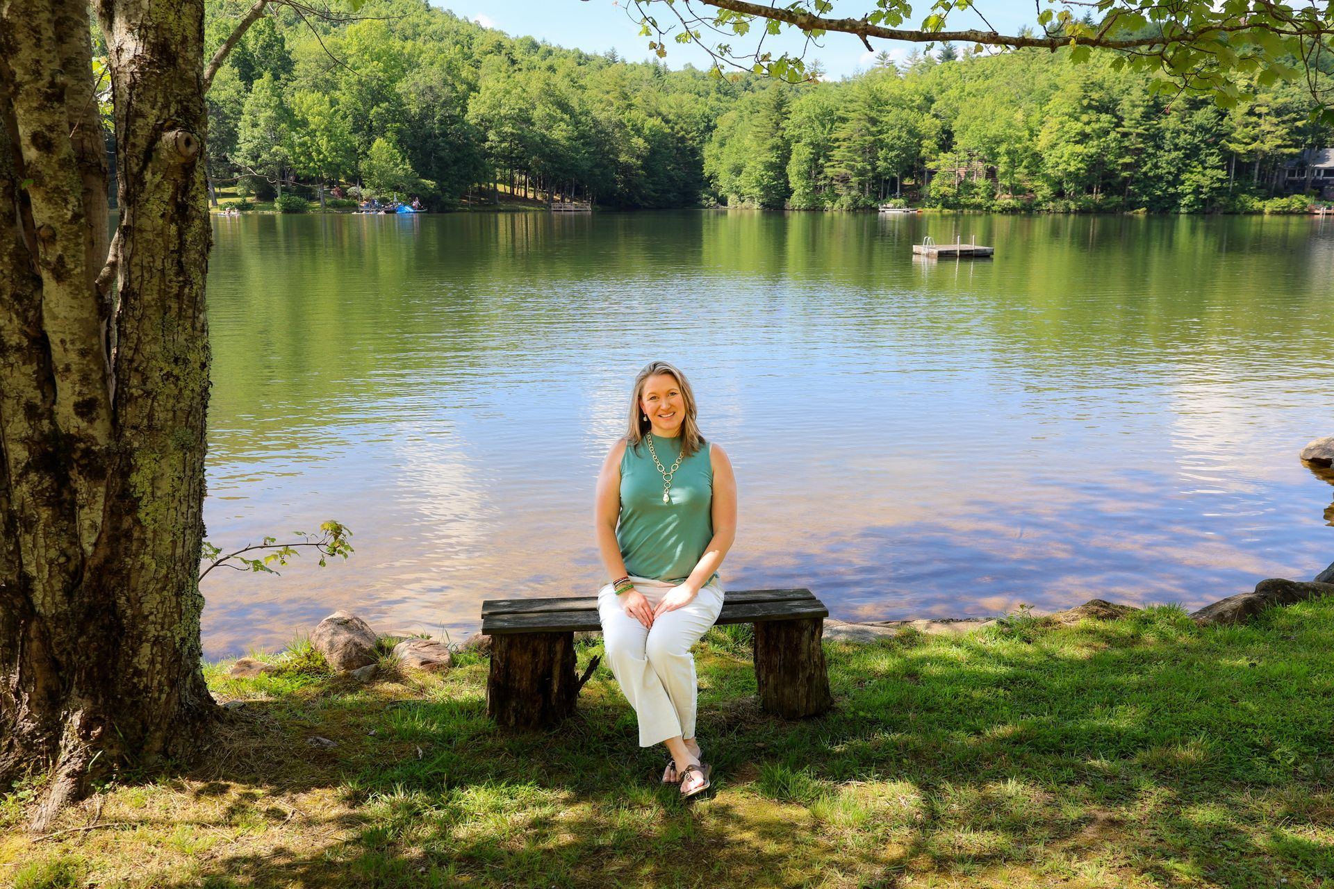 Woman seated on a rustic bench by a lake, smiling. Green top, white pants, trees in background, sunny day.