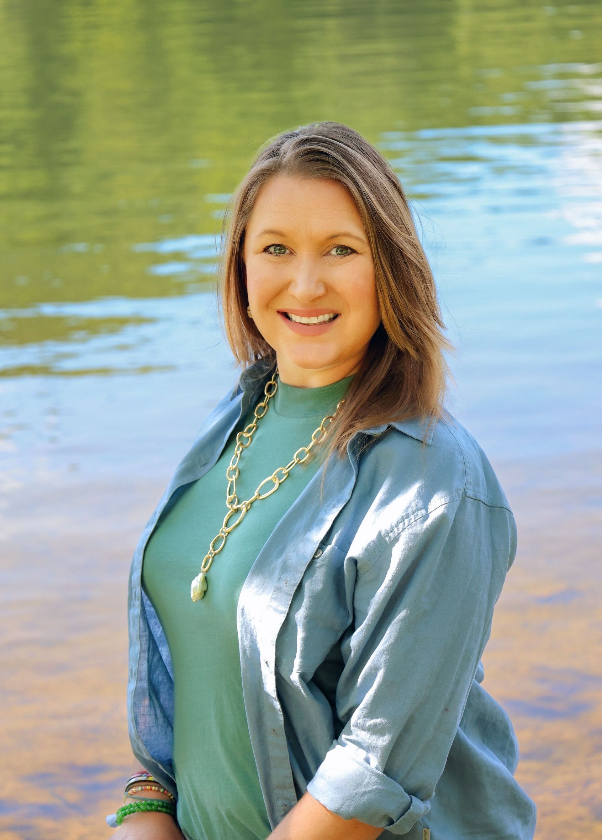 Woman in teal shirt and jacket smiles by a lake, wearing gold necklace and bracelets.
