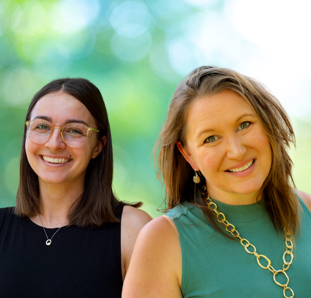 Two smiling women pose outside; one wears glasses and a black top, the other a green top and gold jewelry.
