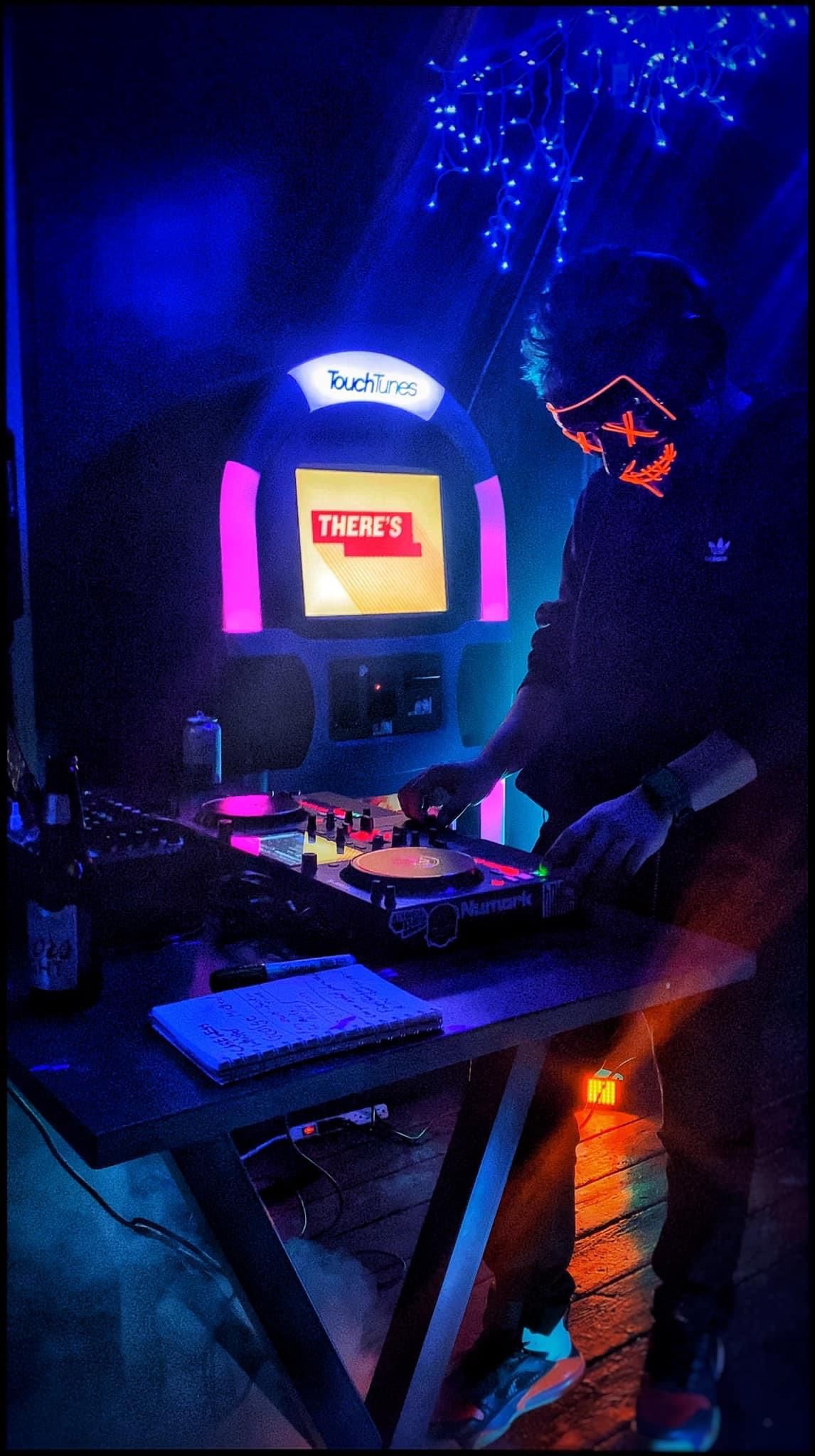 Neon-lit arcade cabinet glowing blue and pink in a dark room, with a table and chair in the foreground